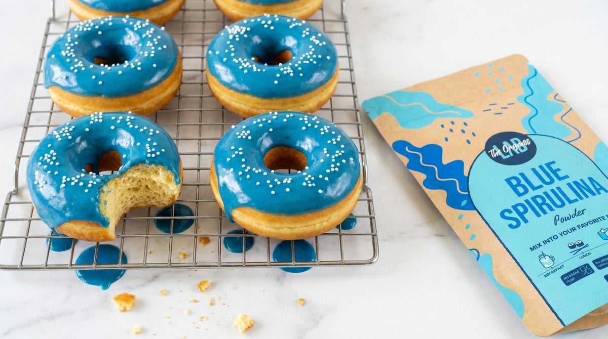 Homemade blue spirulina glazed donuts with glossy blue icing and white sugar sprinkles beside The Organic Labs Blue Spirulina Powder 50g.