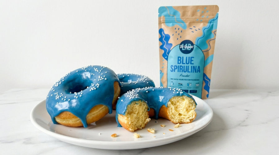 Three blue-glazed spiral donuts on a white plate with a partially eaten donut showing the golden interior, next to a stand-up pouch of HAD Blue Spirulina powder on a light marble surface against a white background.