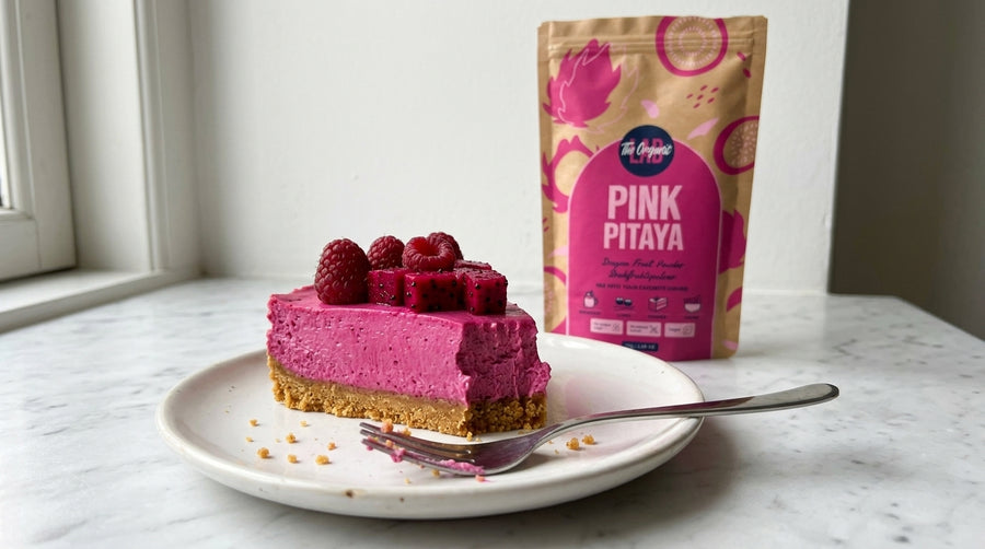 A slice of pink berry cheesecake on a white plate with a fork, standing next to a stand-up pouch labeled 'Pink Pitaya' on a light marble surface near a window.
