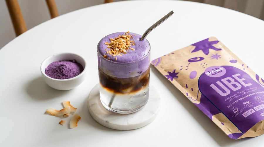 A glass of iced coffee topped with thick purple ube paste and crushed nuts, set on a white table next to a small bowl of purple powder and a kraft pouch labeled ube.