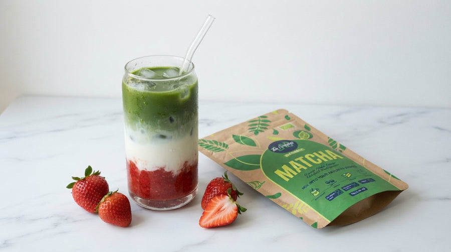 A glass of layered matcha iced drink with green, white, and red layers, shown next to a brown and green matcha pouch on a white marble surface with fresh strawberries.