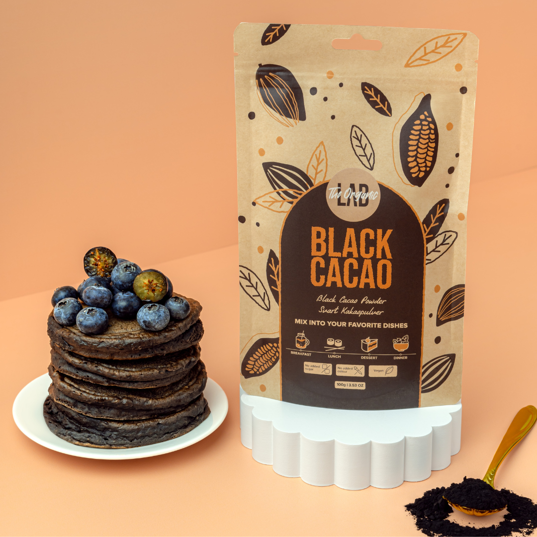 The Organic Lab Black Cacao Powder 100 g pouch next to a stack of black cacao pancakes topped with blueberries